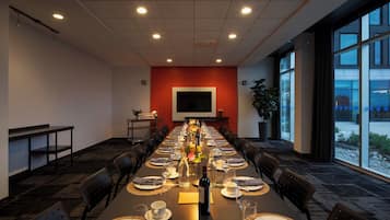 A conference room with a long table set for a meeting, equipped with chairs, a projector screen, and a potted plant.