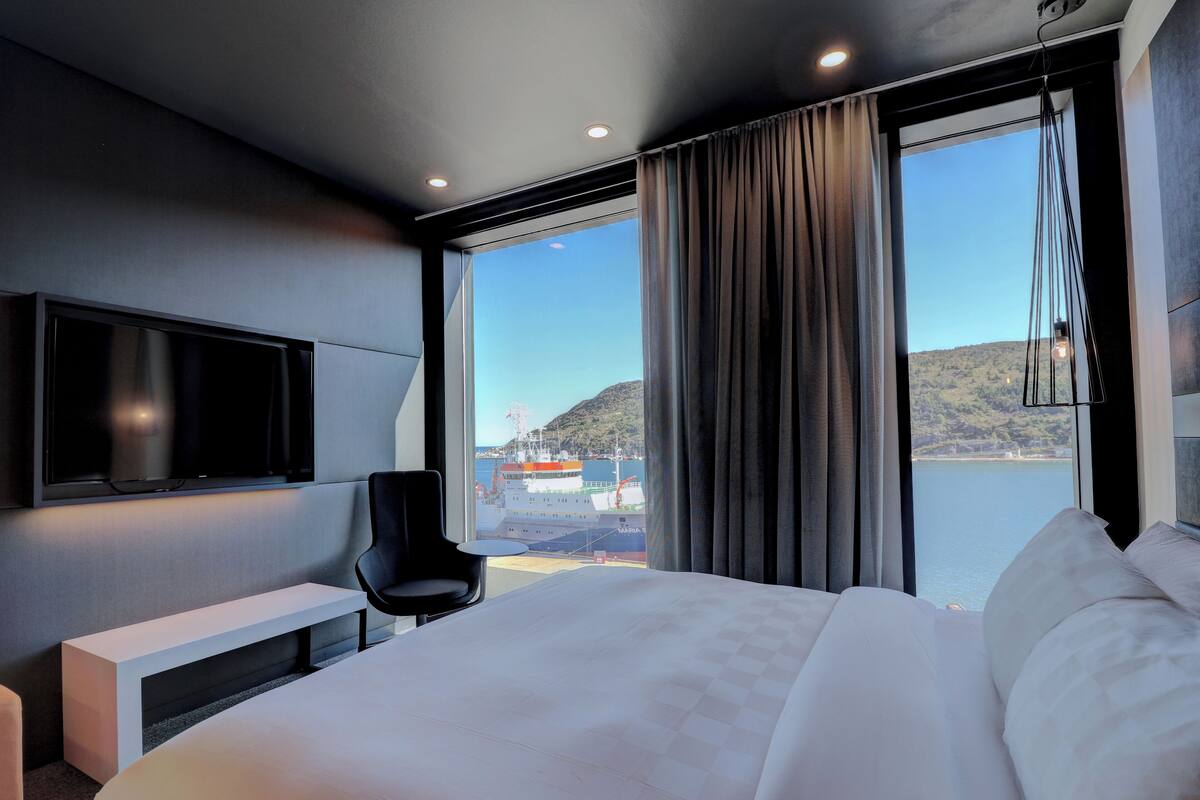A modern hotel room with a large bed, a TV mounted on the wall, a chair, and a large window offering a view of the sea and a ship.