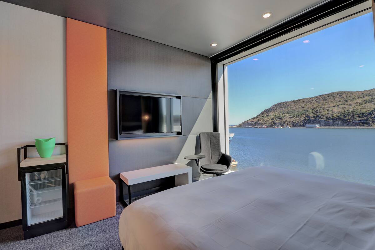 A modern hotel room with a large bed, a television, a small table, a chair, and a view of a body of water and a hilly landscape.