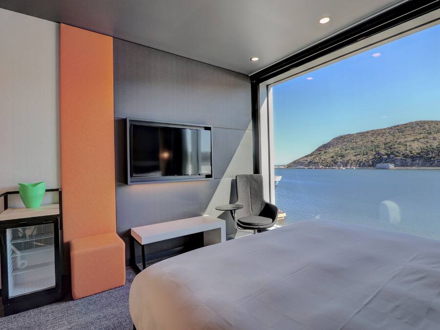 A modern hotel room with a large bed, a television, a small table, a chair, and a view of a body of water and a hilly landscape.