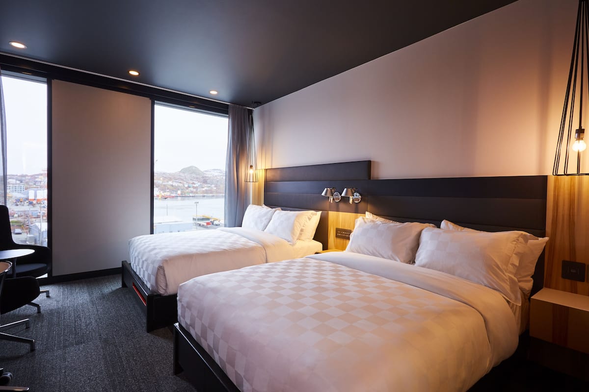 A hotel room with two beds, a desk, and a large window offering a city view.