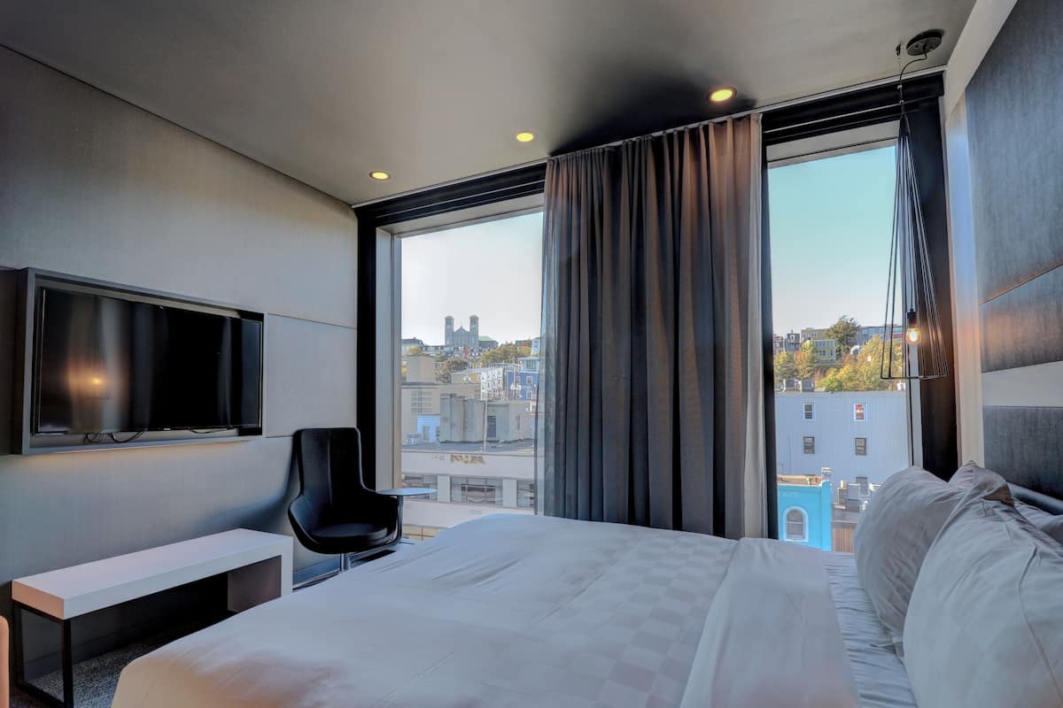 A modern hotel room with a large bed, a flat-screen TV mounted on the wall, a desk, and a view of the cityscape through large windows.