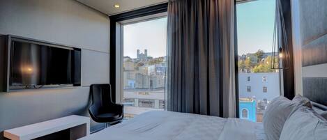 A modern hotel room with a large bed, a flat-screen TV mounted on the wall, a desk, and a view of the cityscape through large windows.