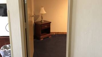 Una habitación con alfombra oscura, una mesita de noche de madera con lámpara, y una puerta que da a otra habitación.