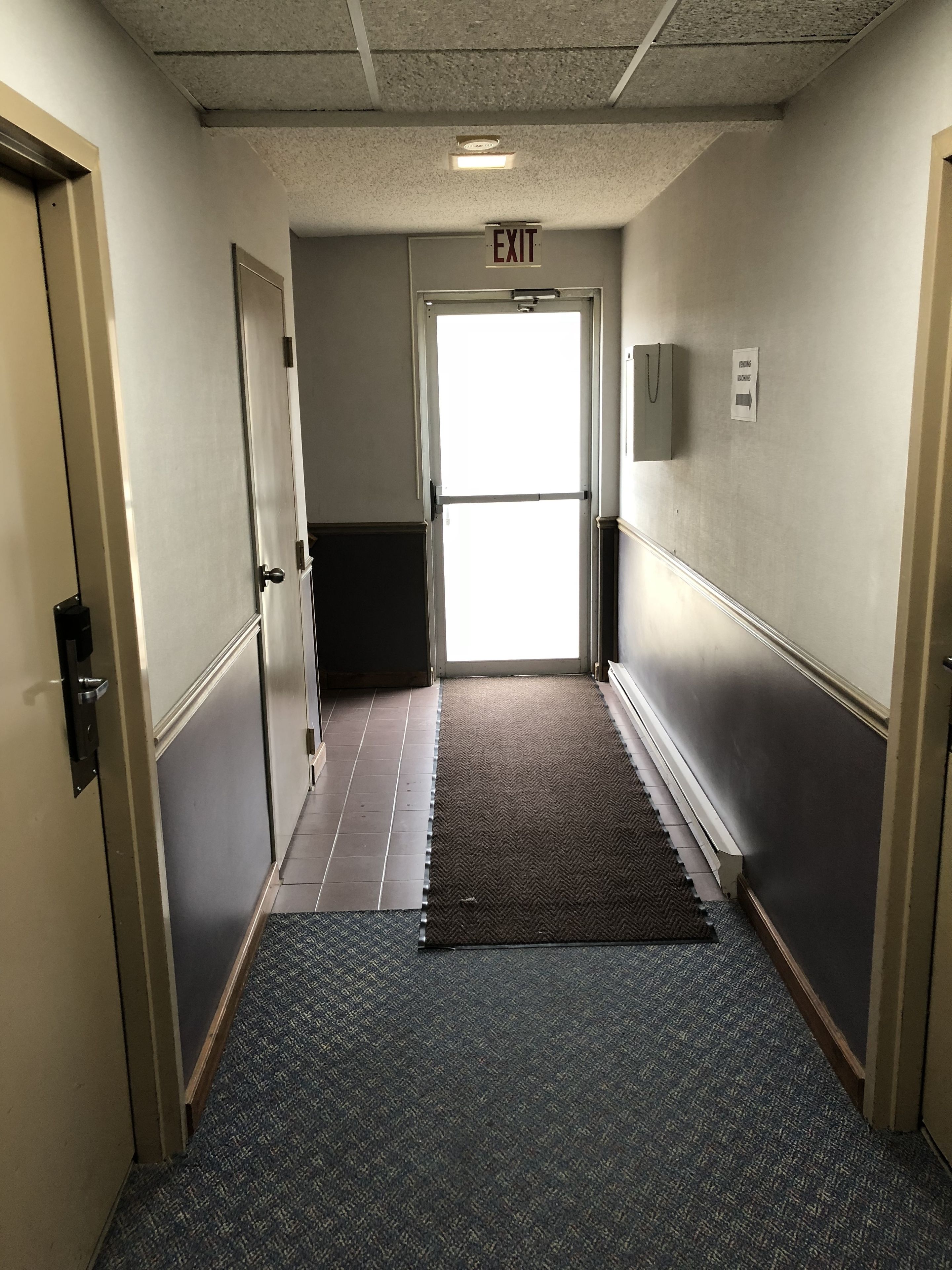 Un pasillo con una señal de salida, una puerta y suelo alfombrado.