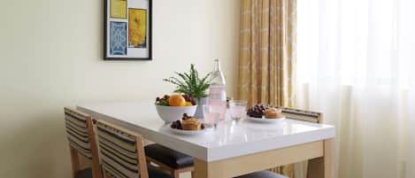 Una mesa de comedor con superficie blanca, rodeada de sillas de madera con cojines negros. En la mesa, hay un cuenco de fruta, una botella y un vaso.