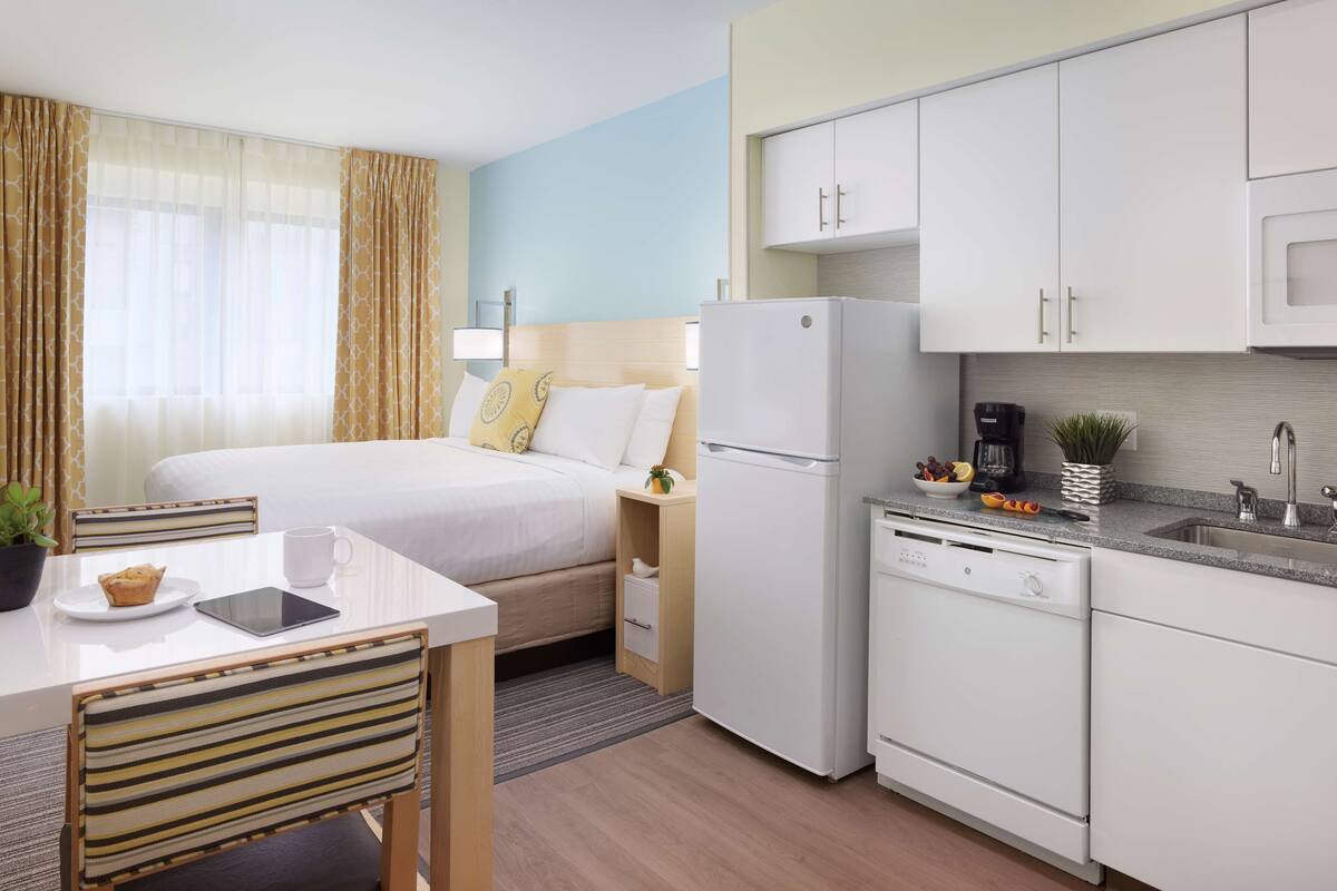 Habitación de hotel compacta con cama, mesita de noche, silla y un área de kitchenette que incluye refrigerador, microondas y pileta.
