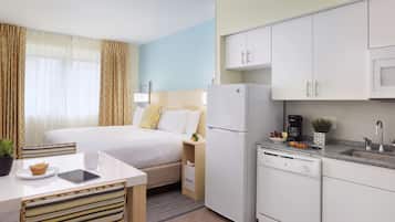 A compact hotel room with a bed, bedside table, chair, and a kitchenette area including a refrigerator, microwave, and sink.