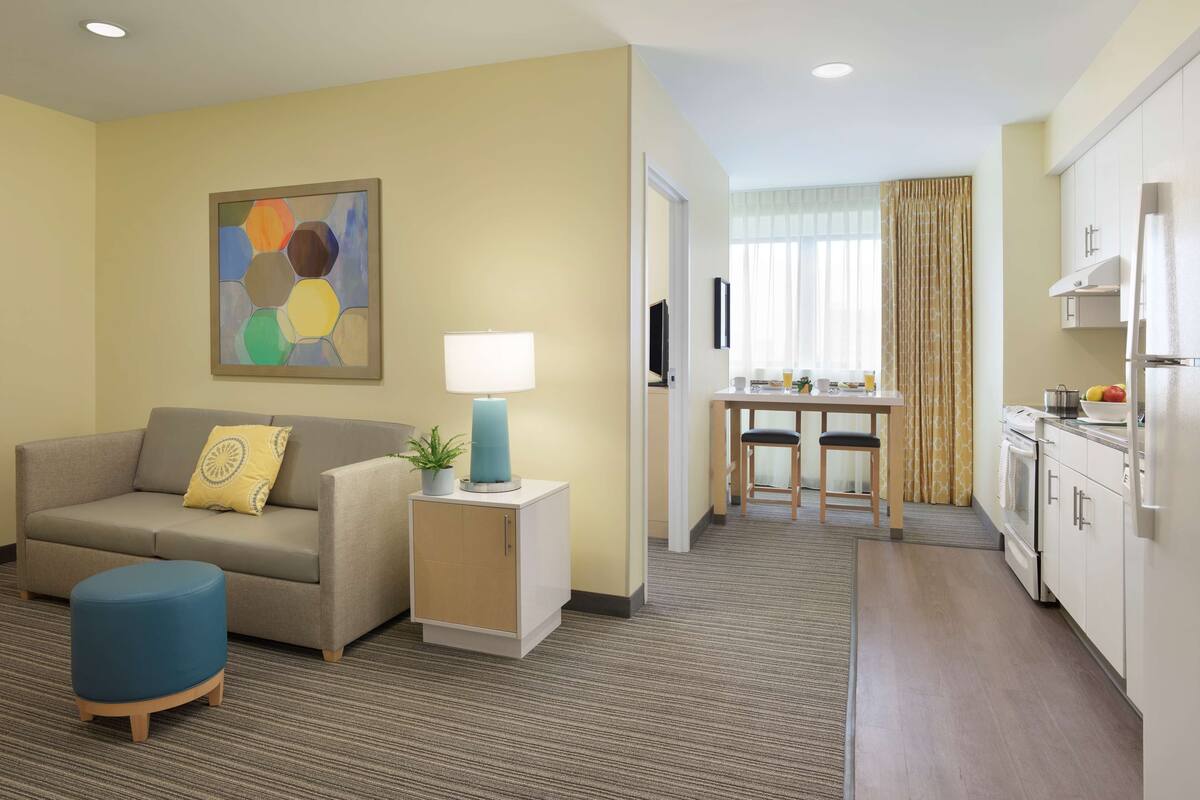 Habitación de hotel compacta con sofá, una pequeña mesa con lámpara, kitchenette y zona de comedor con vistas al exterior.