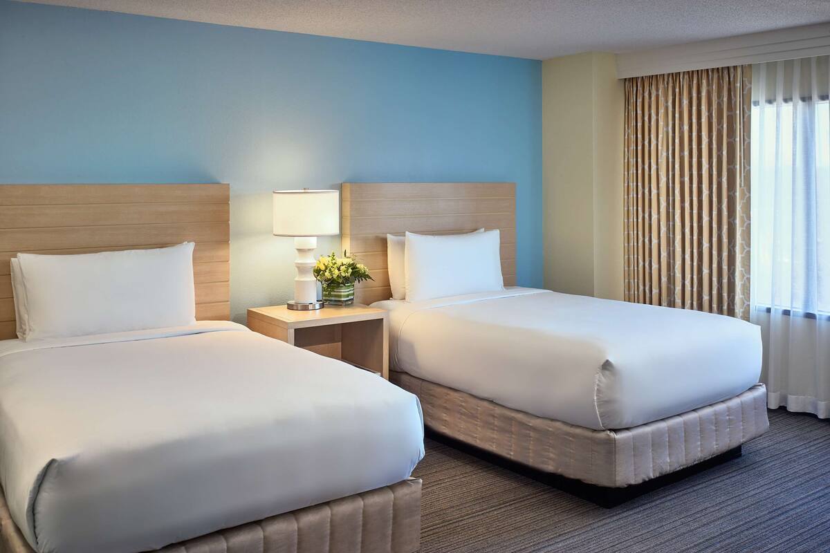 Habitación de hotel con dos camas, una mesita de noche con lámpara y flores, una ventana con cortinas y una pared azul.