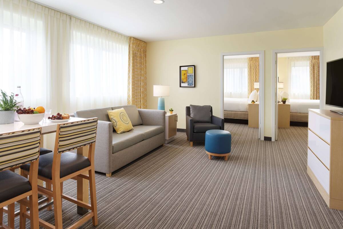 Habitación de hotel con comedor, sofá, sillón, pequeño reposapiés, televisor, lámpara, cama y ventana con cortinas.