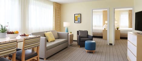 A hotel room with a dining area, a sofa, a chair, a small ottoman, a TV, a lamp, a bed, and a window with curtains.