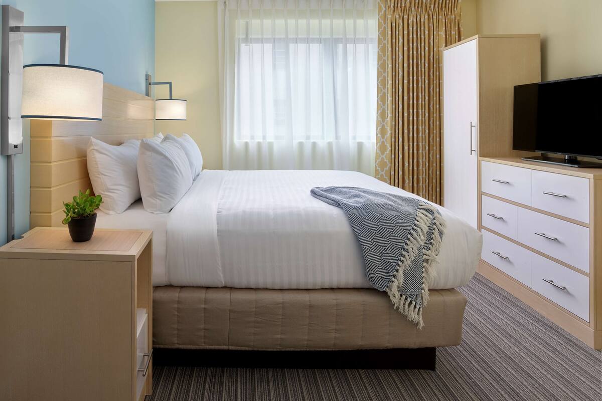 Habitación de hotel con cama, mesitas de noche, televisor, armario y ventana con cortinas.