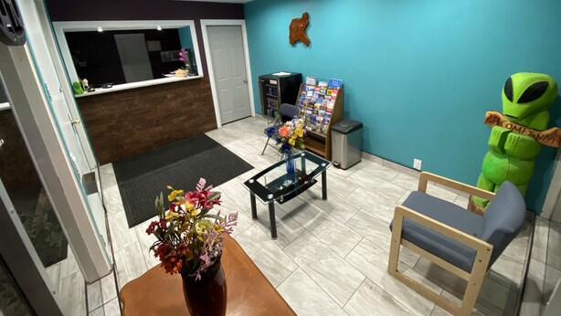 A reception area with a reception desk, a trash can, a chair, a small table with a vase of flowers, and a green alien figure.