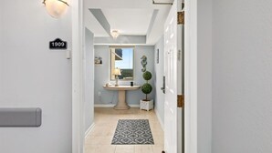 A hallway with a light fixture, a wall-mounted sign reading "1909", a round table with a lamp, a small potted plant, and a patterned rug. - Brighton Tower 1909 3br 3ba Oceanfront 3 Bedroom Condo by RedAwning (Myrtle Beach)