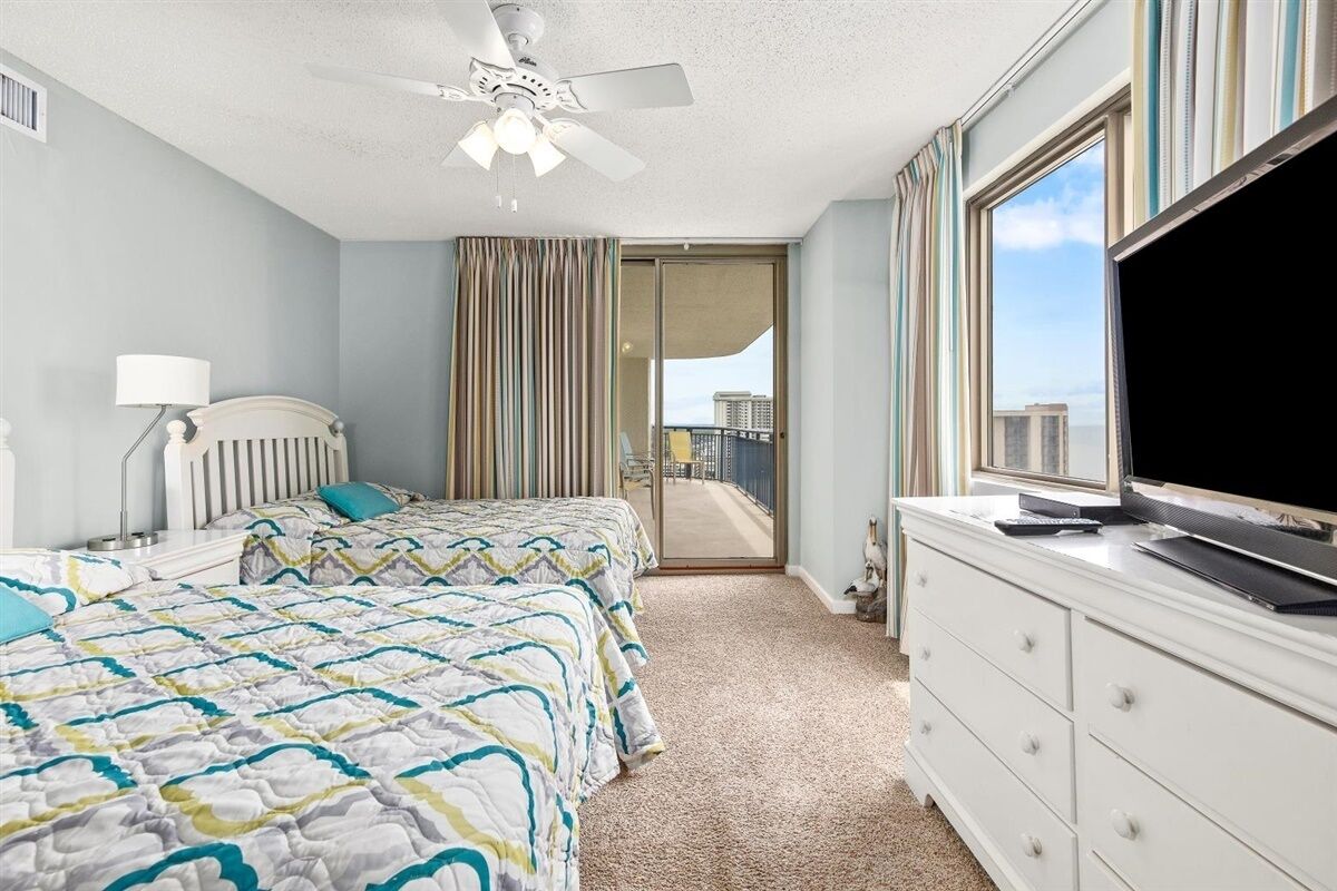 A bedroom with a bed, a ceiling fan, a TV on a dresser, and a view of the outside.