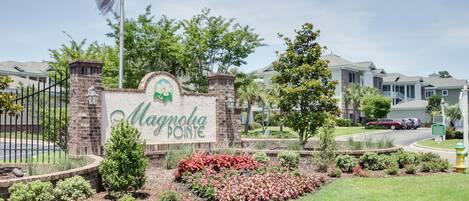 Letrero de Magnolia Pointe con entrada de ladrillo, jardín ajardinado y edificios residenciales al fondo.