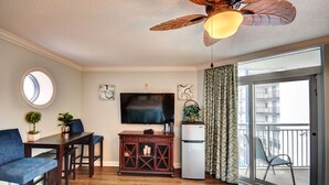 Ein Wohnzimmer mit Deckenventilator, Fernseher, kleinem Kühlschrank, Esstisch mit Stühlen und Blick auf einen Balkon.