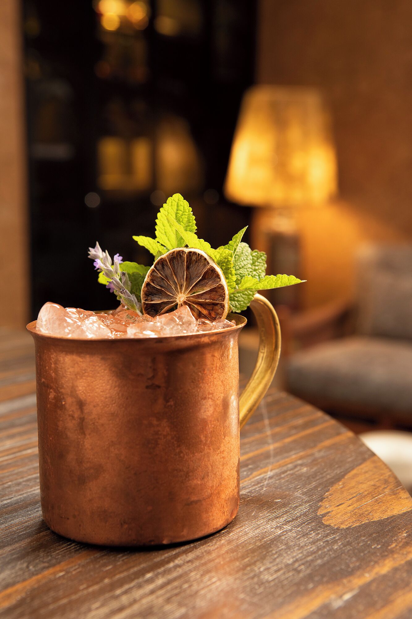 A copper mug filled with a beverage, garnished with mint leaves, a dried orange slice, and a purple flower, placed on a wooden table.