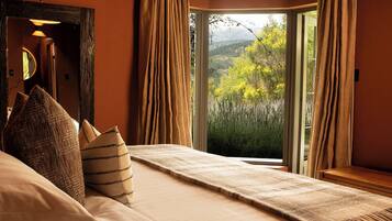 A bedroom with a large bed, wooden beams, a view of greenery, and a window with curtains.