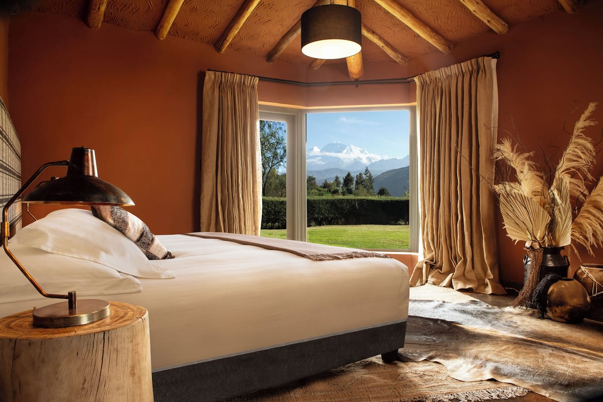 Un dormitorio con cama, mesita de noche, lámpara, ventana con cortinas y vistas a las montañas.
