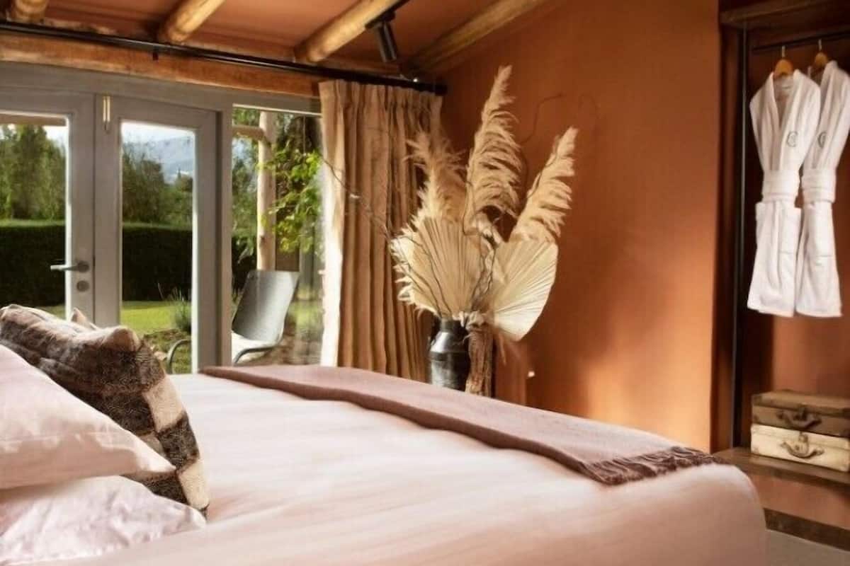 Un dormitorio con cama, una ventana con cortinas, un florero con plumas de pampa y un perchero con batas.