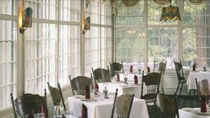 A dining area with multiple tables set for service, large windows, and a view of greenery outside.