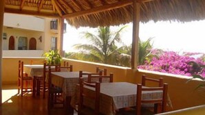A restaurant with wooden tables and chairs, a thatched roof, and a view of purple flowers and palm trees. - Hotel Kootznoowoo (Puerto Escondido)