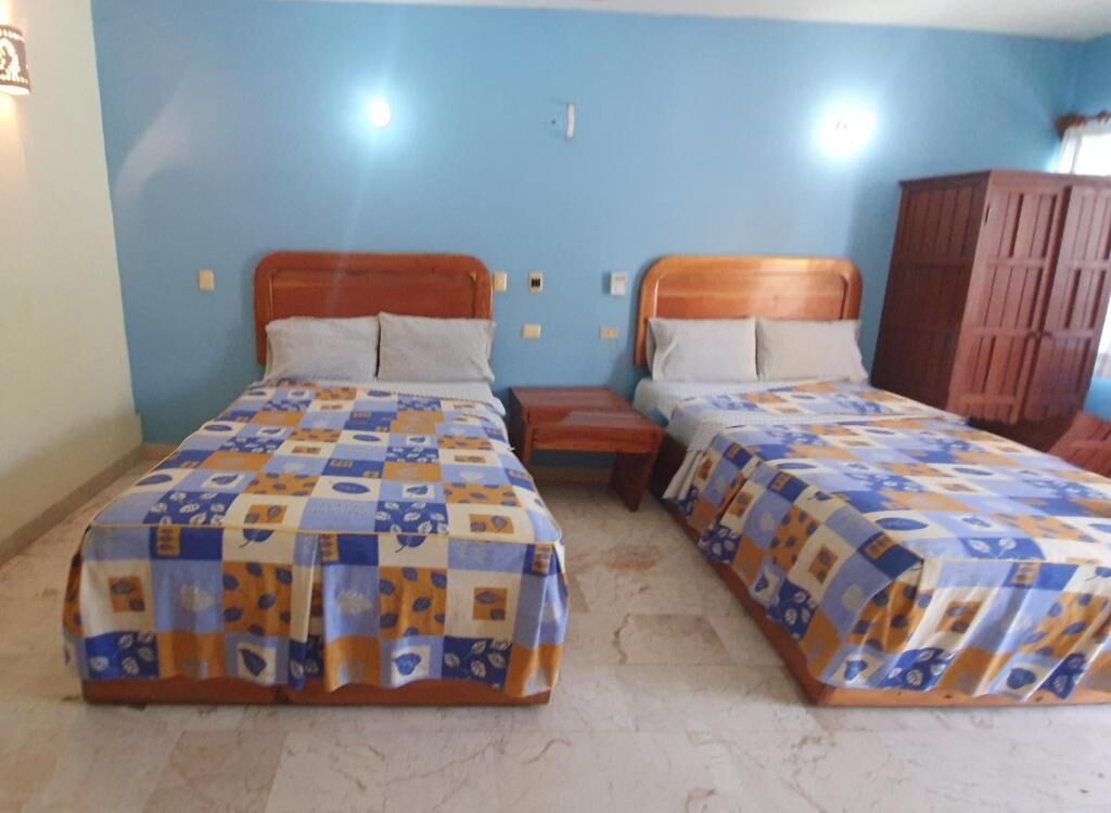 A room with two beds, each with a wooden headboard and a patterned bedspread. There is a wooden nightstand between the beds, and a wooden wardrobe is visible in the corner.