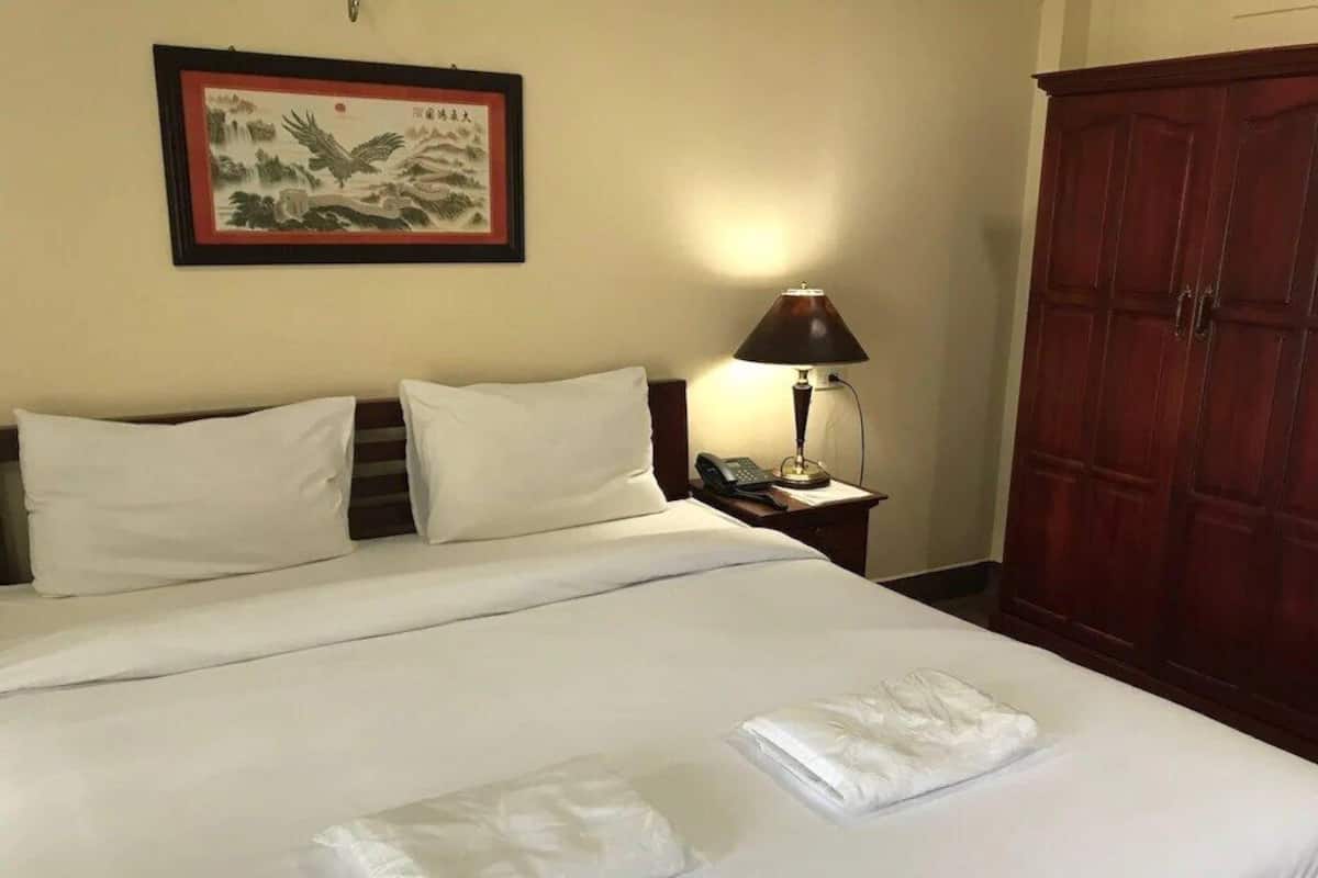 A neatly made bed with white linens, a bedside lamp, a telephone, and a traditional wooden wardrobe.