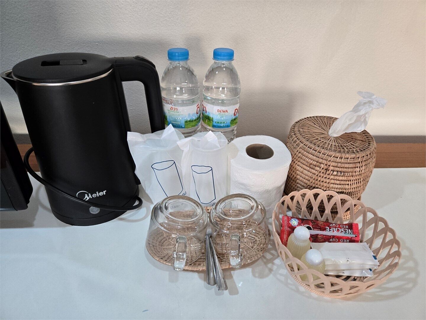 Una mesa con una cafetera de color negro, dos botellas de agua, una cesta con pañuelos y un repuesto, dos tazas de vidrio, una jarra de vidrio y una caja de pañuelos.