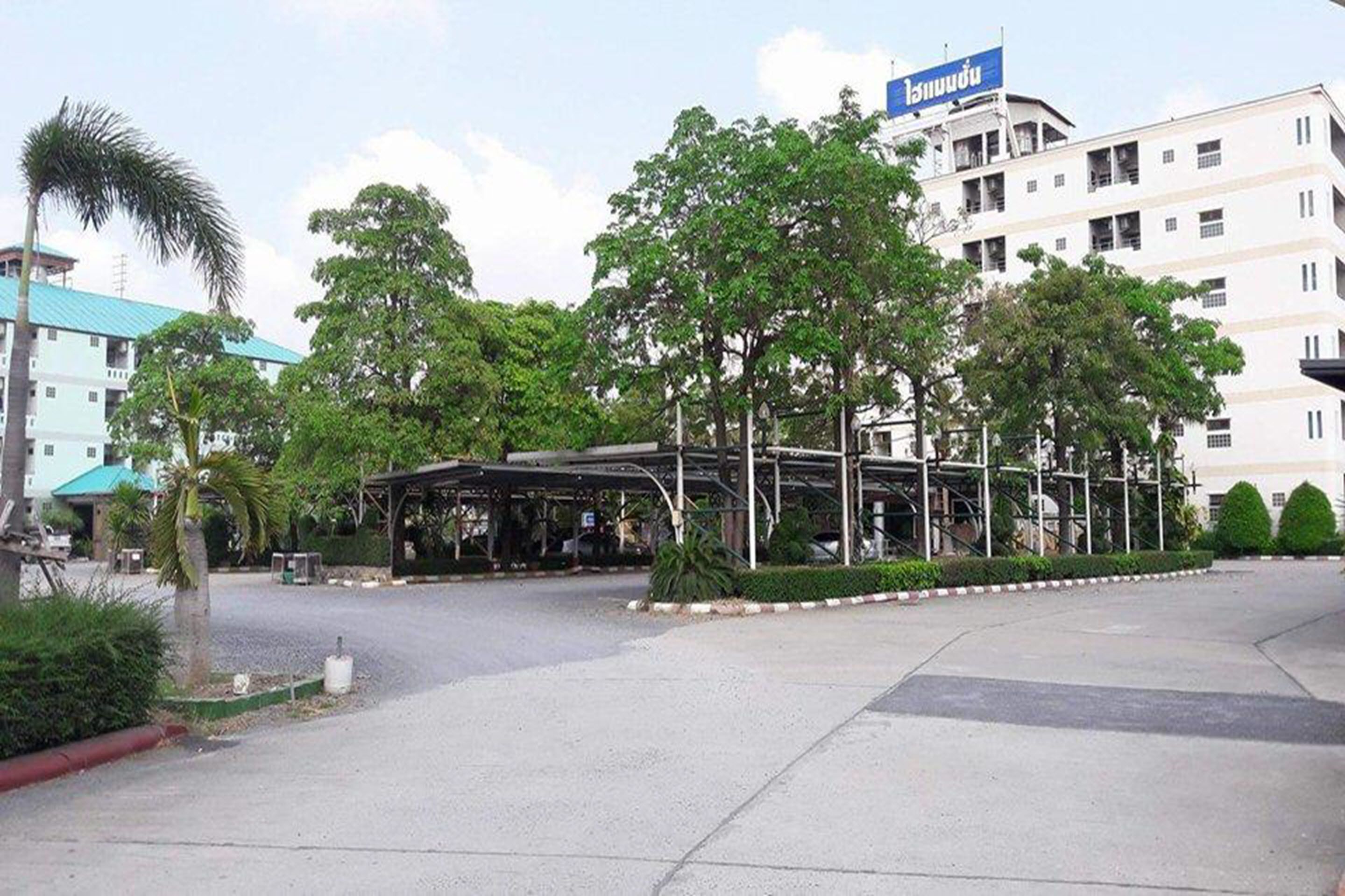 Entrada de hotel con zona de espera cubierta, palmeras y un edificio con un letrero azul al fondo.