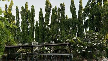 Un área de estacionamiento de concreto con una estructura metálica de puente, rodeada de árboles altos y algunas plantas en macetas.