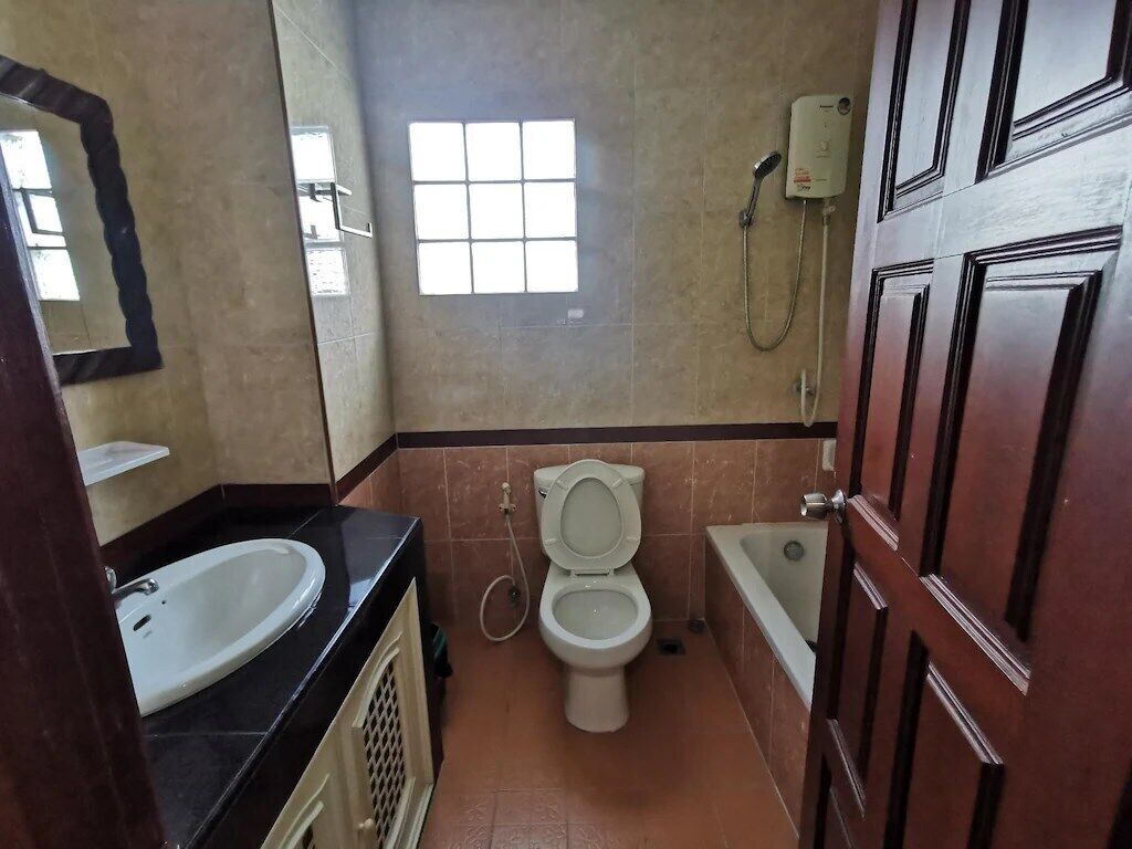 Un baño con inodoro, lavamanos y bañera. Hay una ventana sobre el inodoro y un cabezal de ducha montado en la pared.