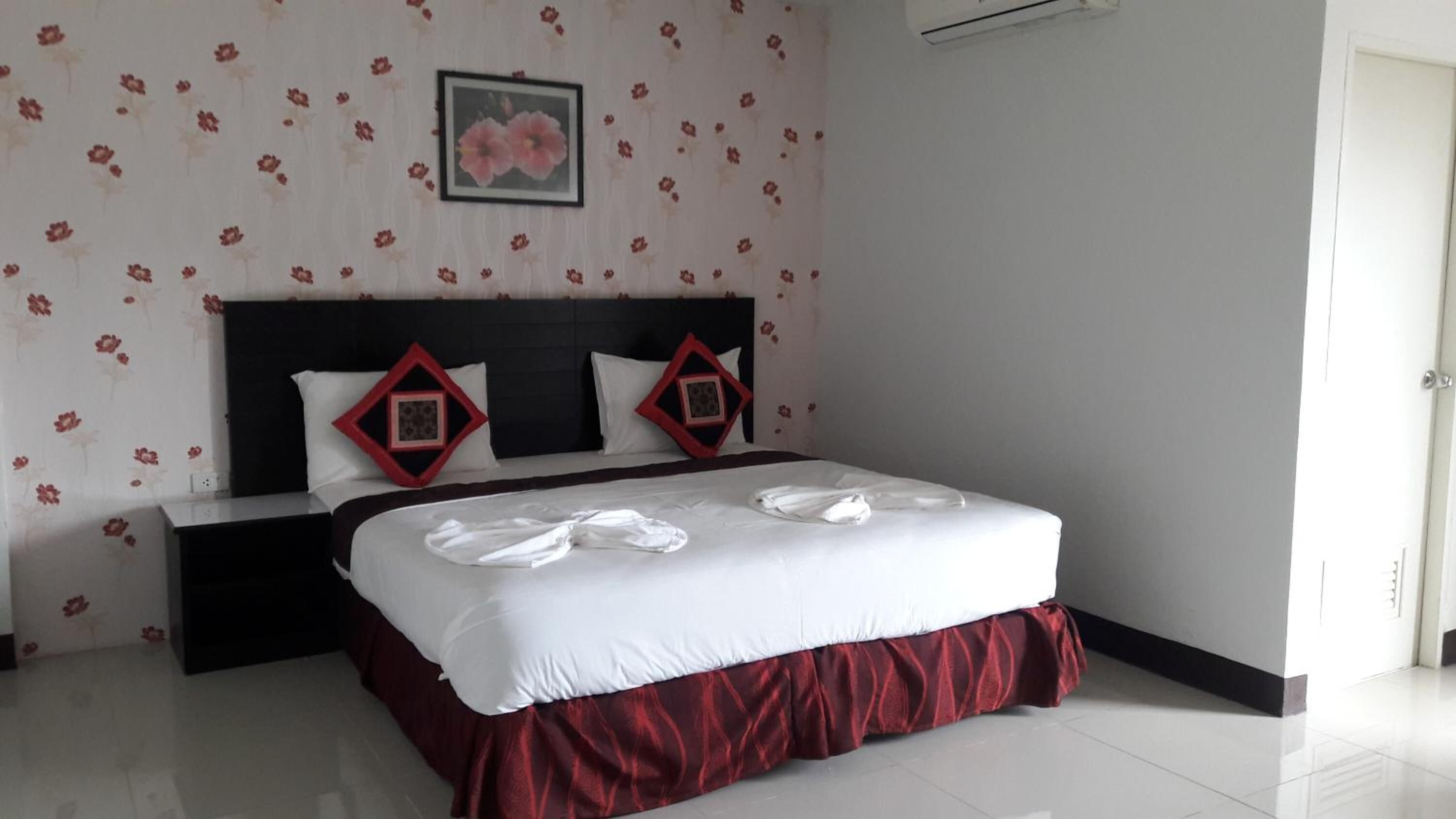 Una cama bien tendida con una colcha blanca y roja, dos almohadas con estampados de diamantes rojos y un cuadro con flores en la pared.