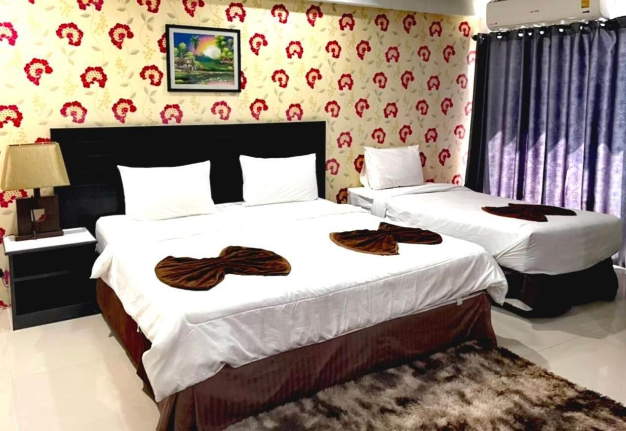 Habitación de hotel con dos camas, una cama grande con cabecera oscura y una cama más pequeña con cabecera blanca. Hay una mesita de noche con una lámpara y un marco de fotos en la pared.