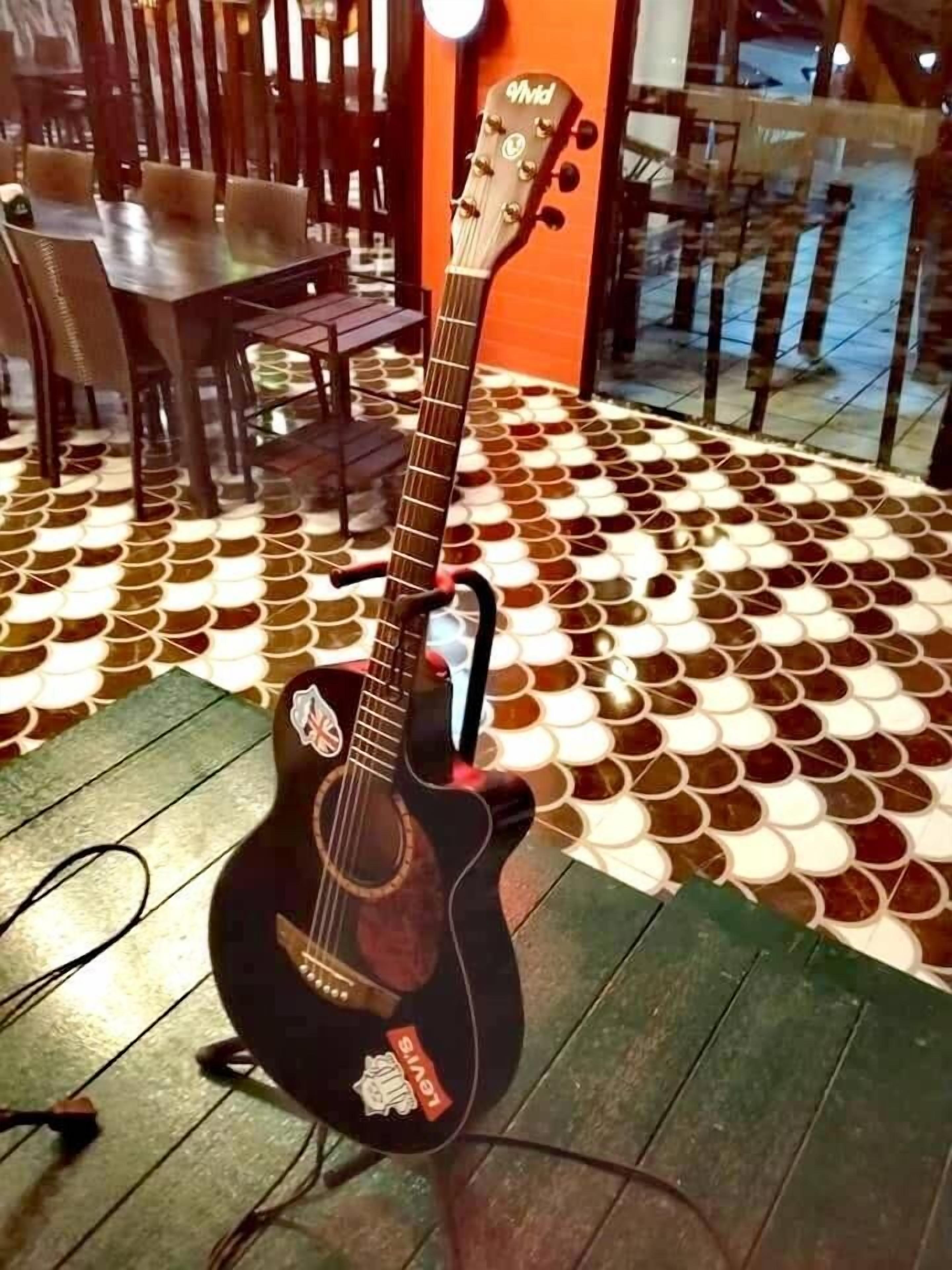 Una guitarra sobre una mesa, con un suelo estampado y sillas al fondo.