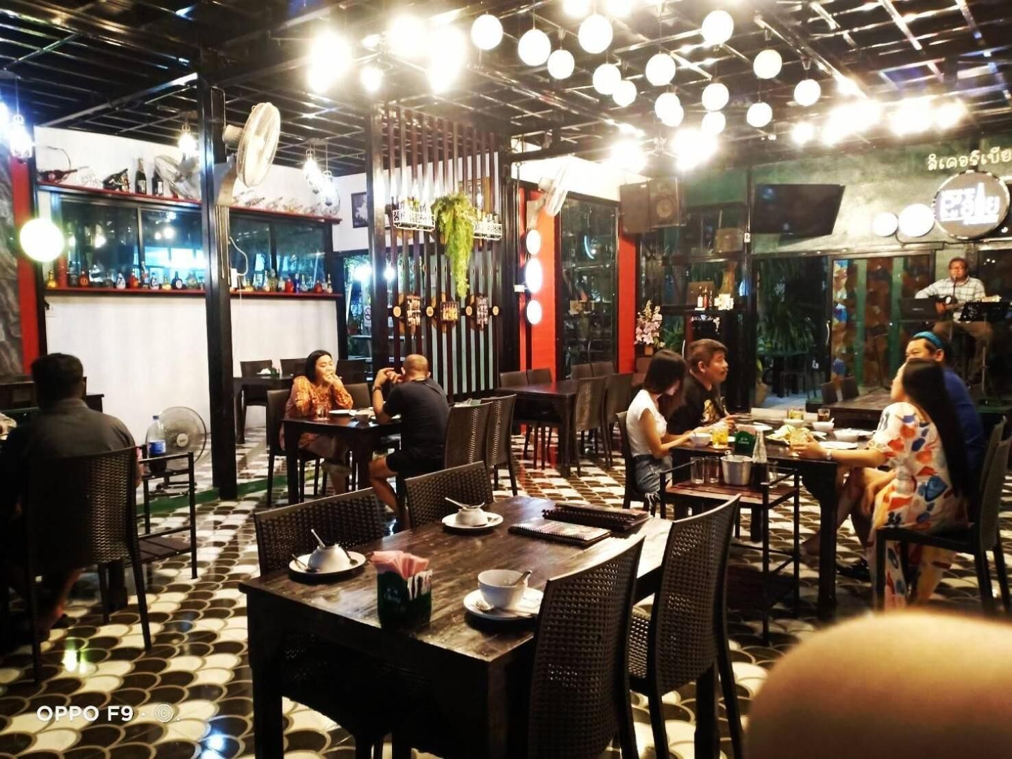 A restaurant with multiple tables and chairs, a bar area with bottles and glasses, and a ceiling with hanging lights.