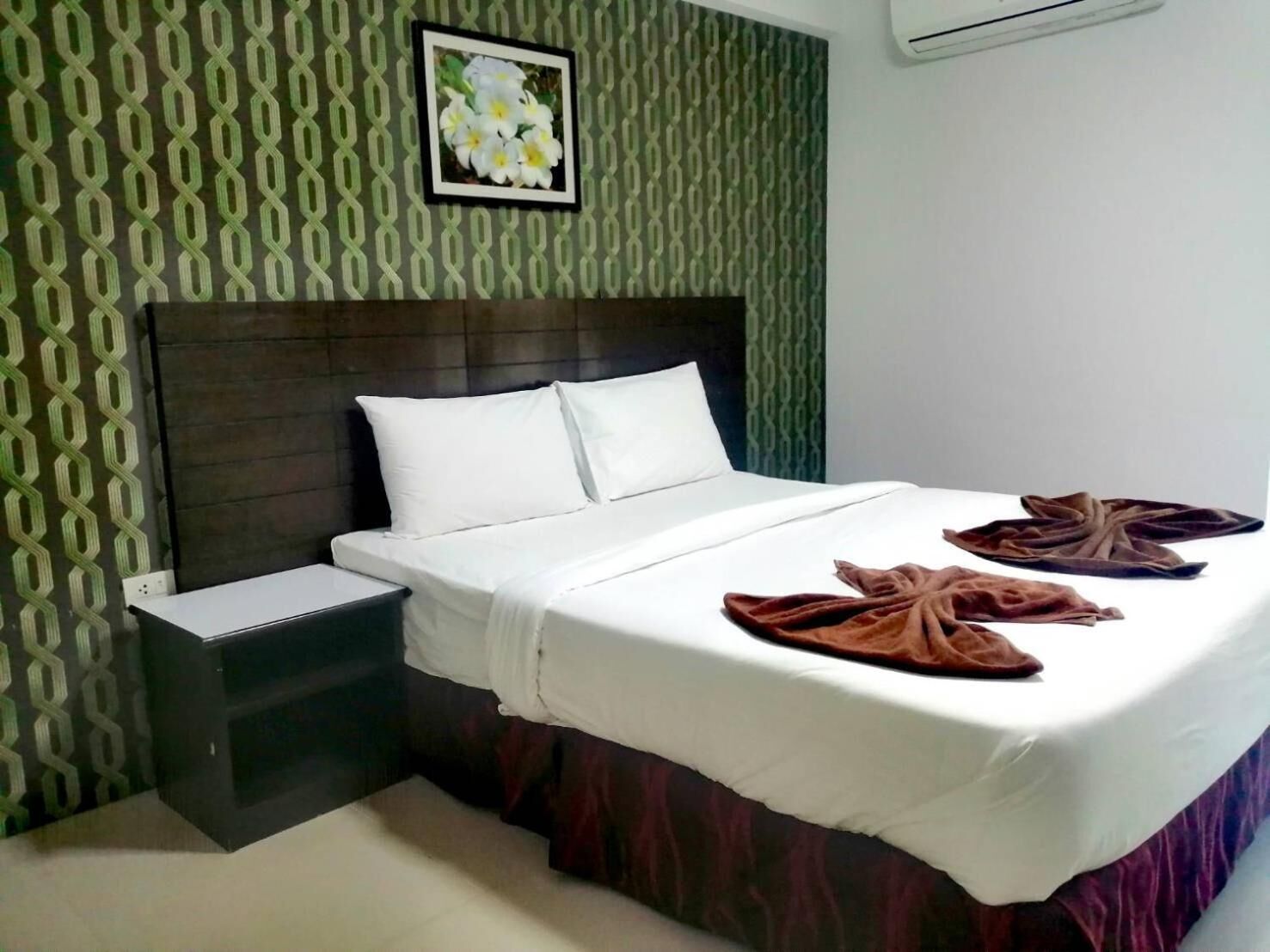 A hotel room with a bed, white linens, a headboard, a nightstand, a wall with a green pattern, and a framed picture above the bed.