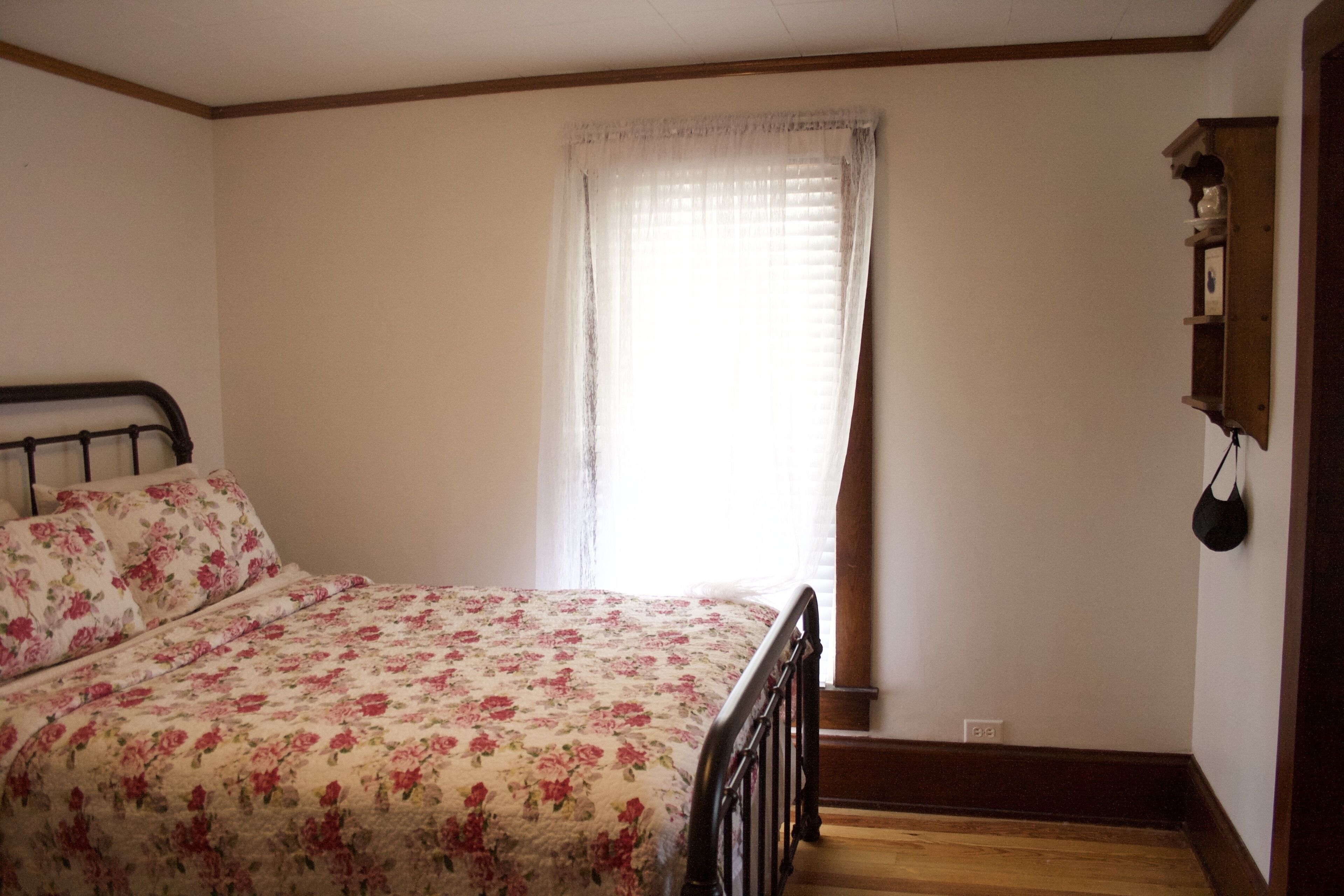 Un dormitorio con colcha estampada, cabecera de madera, ventana con cortinas de gasa y estante de madera con un objeto colgando.