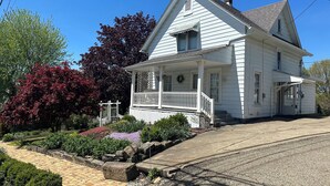 A white two-story house with a porch, a chimney, and a garden with various plants and flowers.