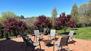 An outdoor patio with brick flooring and metal chairs arranged around a central fire pit.