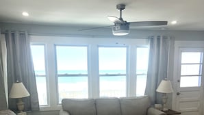 Ein Wohnzimmer mit Sofa, Sessel und Couchtisch, großen Fenstern mit Meerblick und Deckenventilatoren.