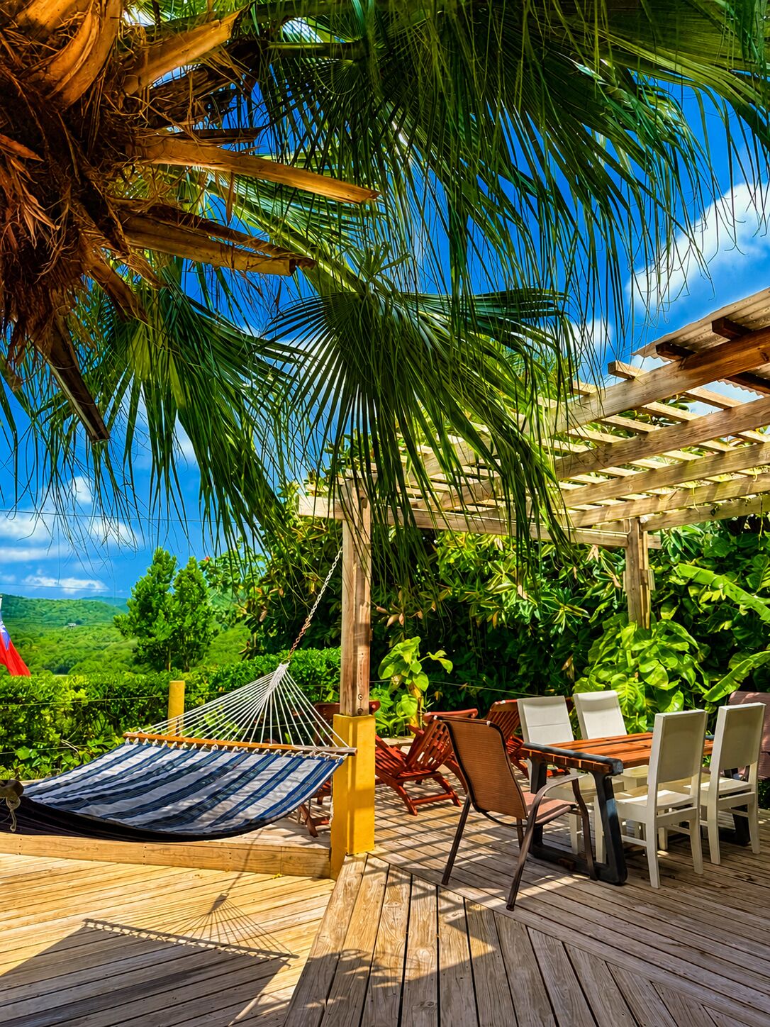 A wooden deck with a hammock, outdoor chairs, and a table, surrounded by lush greenery and a clear sky.