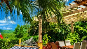 A wooden deck with a hammock, outdoor chairs, and a table, surrounded by lush greenery and a clear sky. - 3 bedroom Villa with Pool + Ocean Views - Overlooks Golf course (Gros Islet)