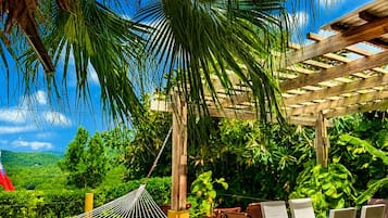 A wooden deck with a hammock, outdoor chairs, and a table, surrounded by lush greenery and a clear sky.