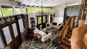 A spacious living area with a stone floor, a sofa set, a wooden staircase, and a stone fireplace. - 3 bedroom Villa with Pool + Ocean Views - Overlooks Golf course (Gros Islet)