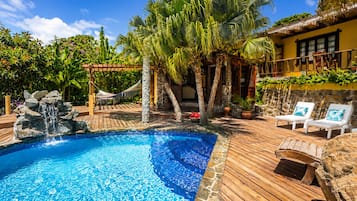 Ein Pool mit Wasserfall, umgeben von Holzterrasse, Palmen und Liegestühlen.