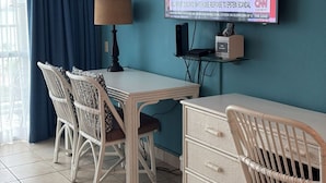 Une chambre d’hôtel avec un mur de couleur turquoise, un téléviseur à écran plat fixé au-dessus d’un bureau en bois, des fauteuils en osier et une fenêtre ornée de rideaux bleus.