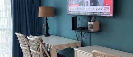 A hotel room with a teal wall, a flat-screen TV mounted above a wooden desk, wicker chairs, and a window with blue curtains.